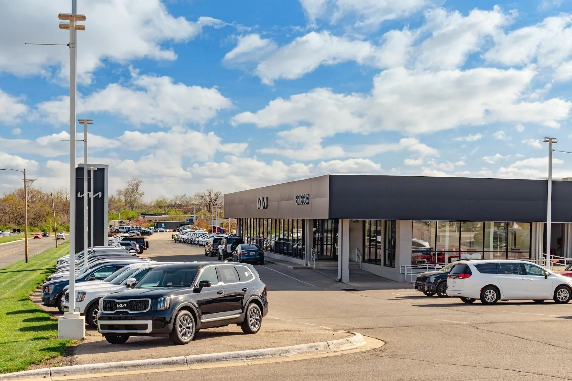 A Kia dealership building with the Kia logo above the showroom glass and new vehicles parked across the lot in front of it.