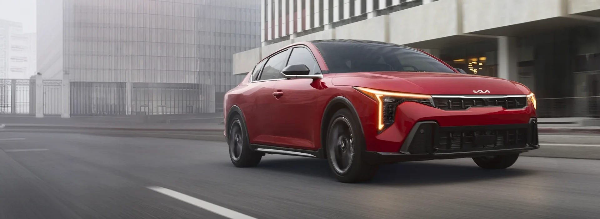 Red 2026 Kia K4 GT-Line Turbo driving along a city street past a glass office building