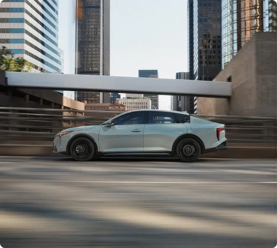 Sage green 2026 Kia K4 shown in profile driving beneath a city overpass, highlighting its fastback roofline