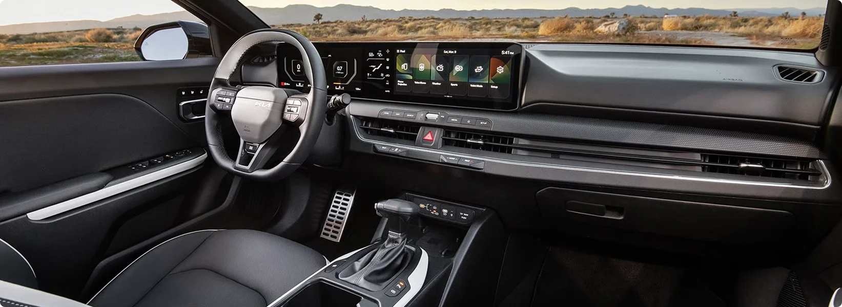 Wide view of the 2026 Kia K4 dashboard with the sport steering wheel and dual panoramic displays, a desert highway ahead through the windshield