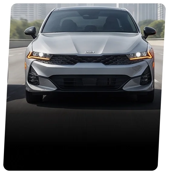 Silver Kia K5 sedan shown head-on with its LED headlights lit on a highway