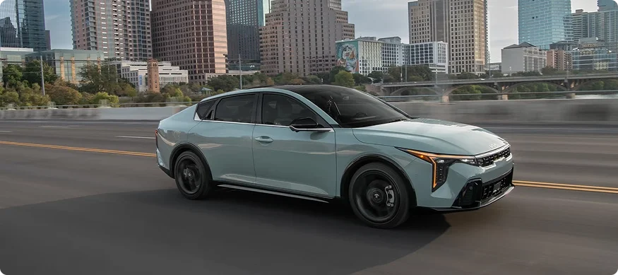 Sage green 2026 Kia K4 driving along a highway with a downtown skyline and river behind it
