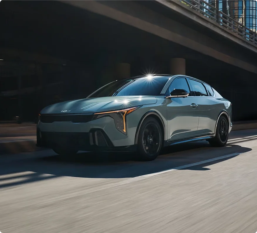 Sage green 2026 Kia K4 with its headlights lit driving beneath a concrete overpass
