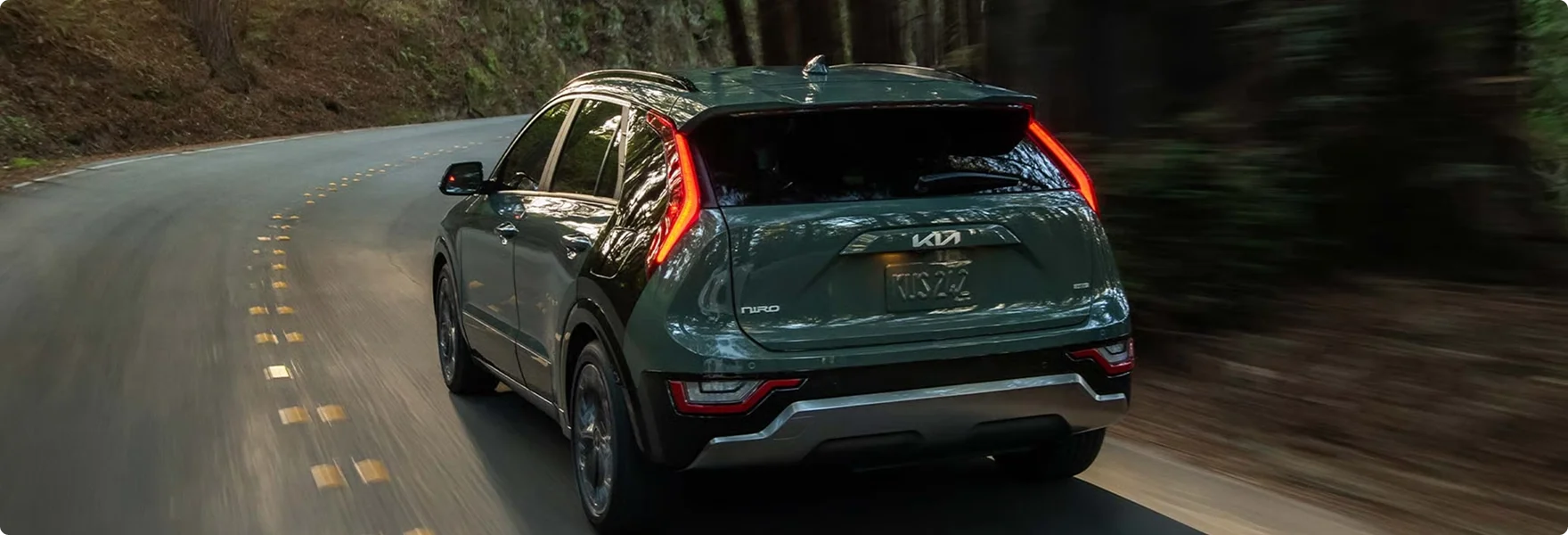Dark green 2026 Kia Niro Hybrid seen from behind as it drives along a winding forest road