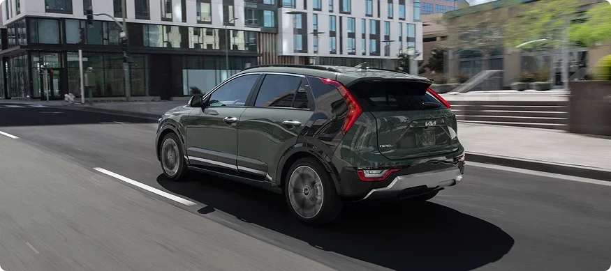 Dark green 2026 Kia Niro Hybrid driving down a city street past modern glass buildings