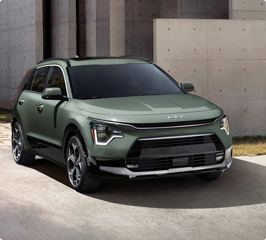Sage green 2026 Kia Niro Hybrid shown from the front three-quarter view beside a poured-concrete wall