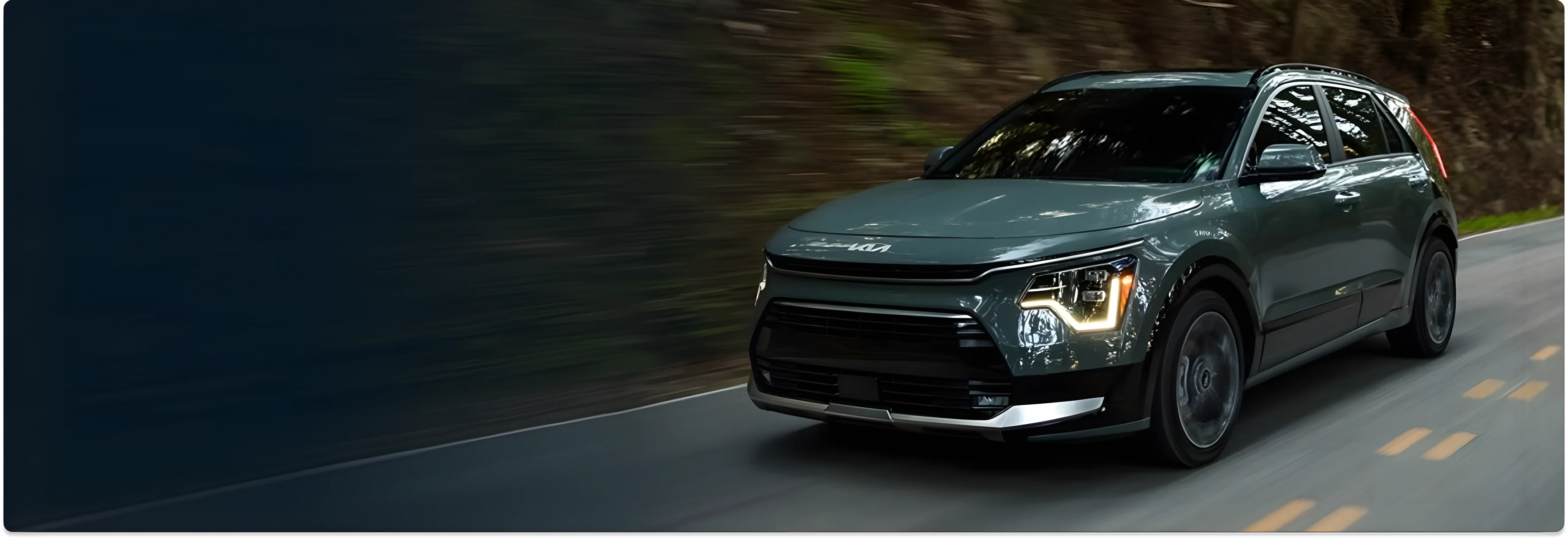 Dark green 2026 Kia Niro Hybrid driving along a tree-lined mountain road
