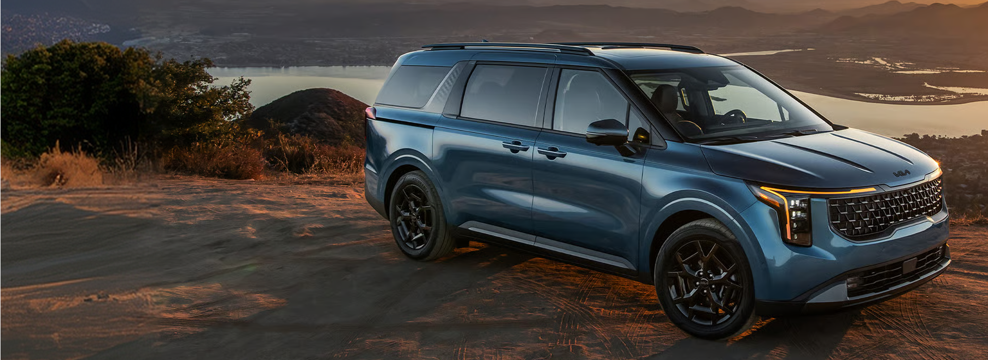 Blue 2027 Kia Carnival MPV parked on a mountain overlook above a lake at sunset