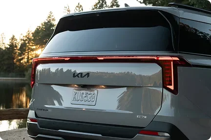 Rear of a grey Carnival MPV Hybrid with illuminated tail lights beside a lake at sunset