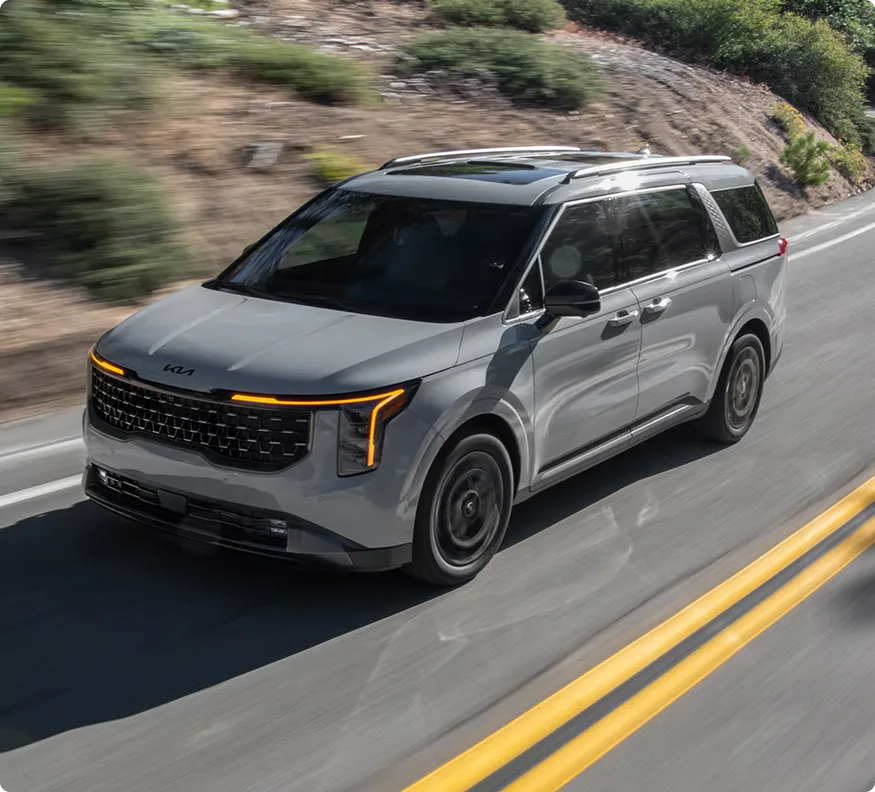 Grey 2027 Carnival MPV Hybrid driving on a winding mountain road, front three-quarter view