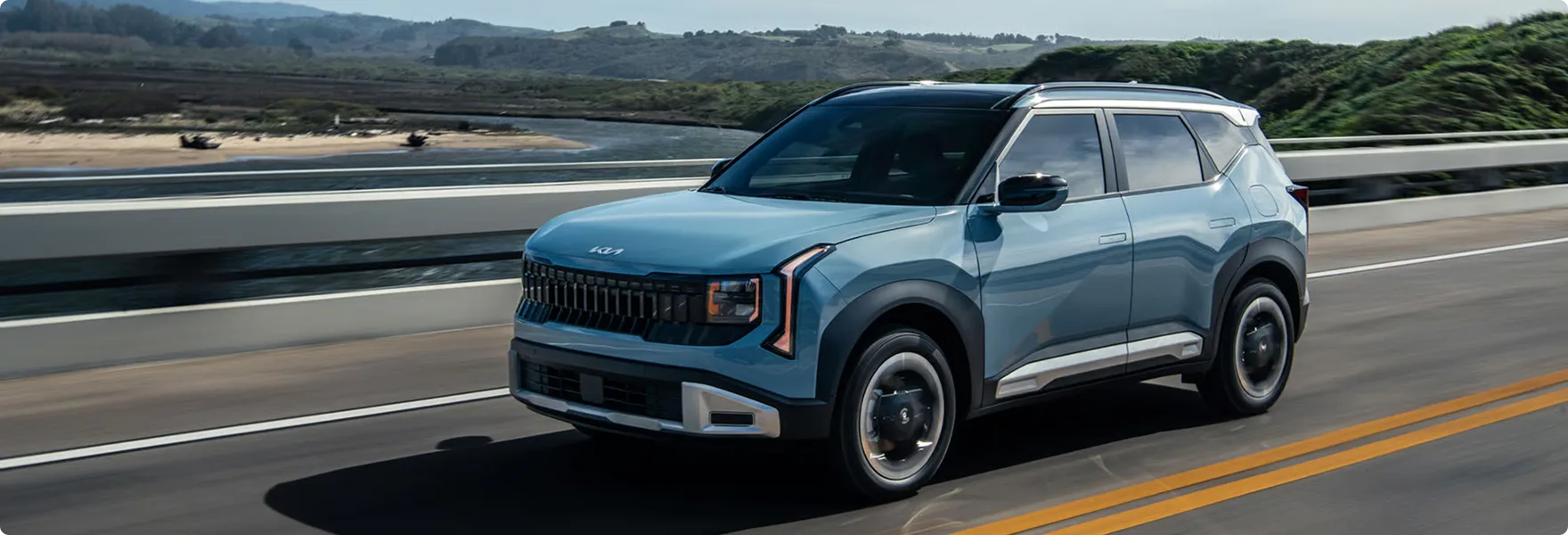 Light blue 2027 Kia Seltos driving on a coastal highway, front three-quarter view