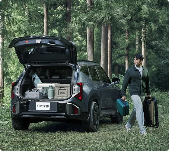 Person carrying gear to the open hands-free power liftgate of a dark green Seltos in a forest
