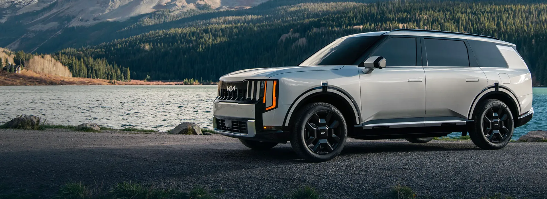Silver 2027 Kia Telluride Turbo-Hybrid parked by a lake with forested mountains behind
