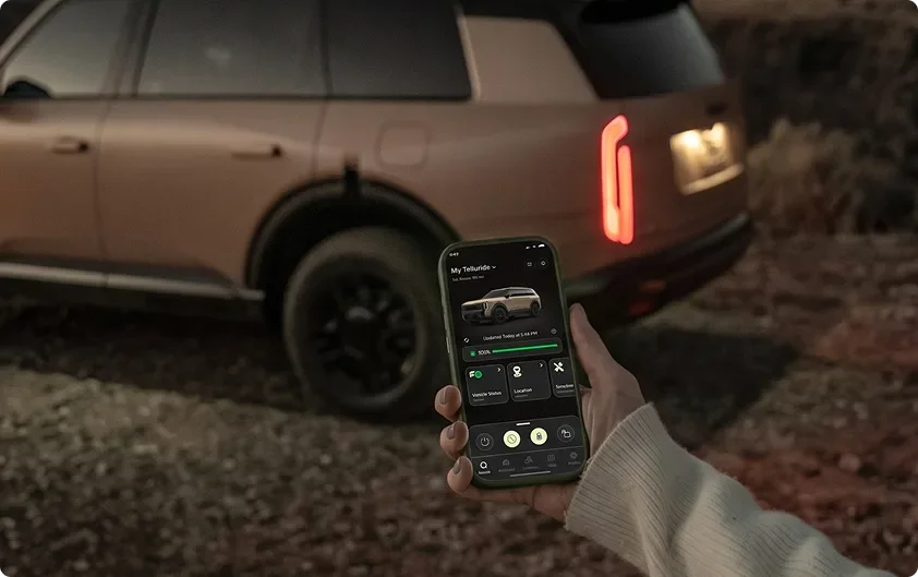Hand holding a smartphone running the Kia app beside a bronze Telluride Hybrid at dusk