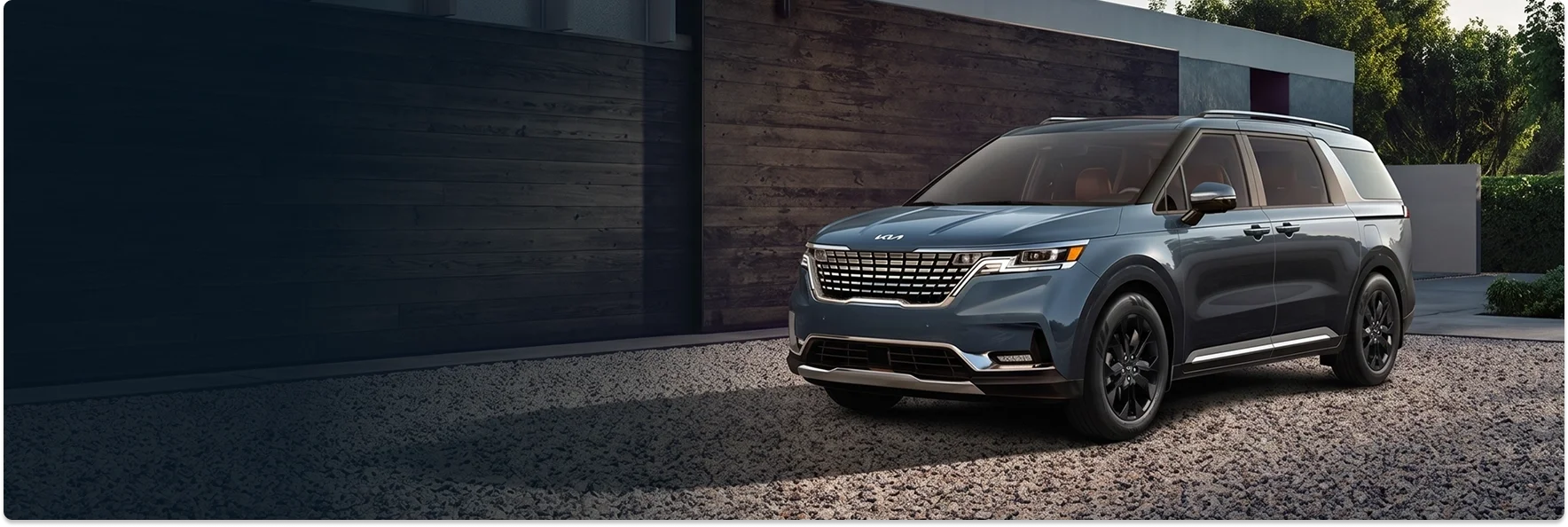 Blue-gray Kia Carnival parked on a gravel drive beside a wood-paneled modern home