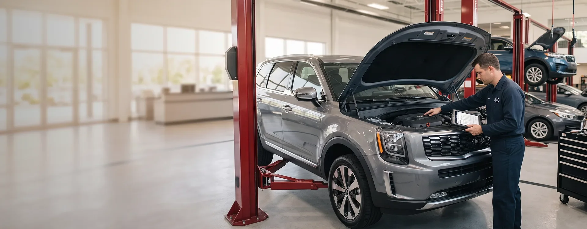 Briggs Kia of Topeka technician reviewing a tablet at the open hood of a silver Kia Telluride raised on a service lift