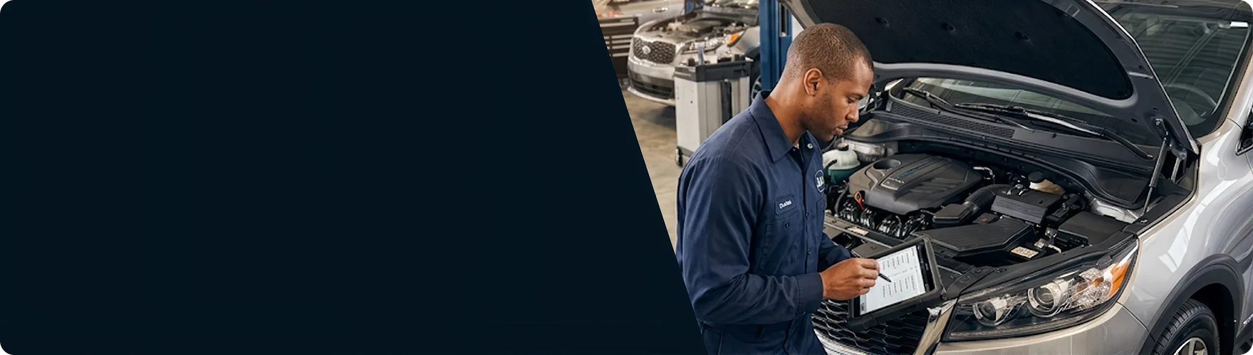 Kia technician logging multipoint inspection results on a tablet at the open hood of a silver Kia